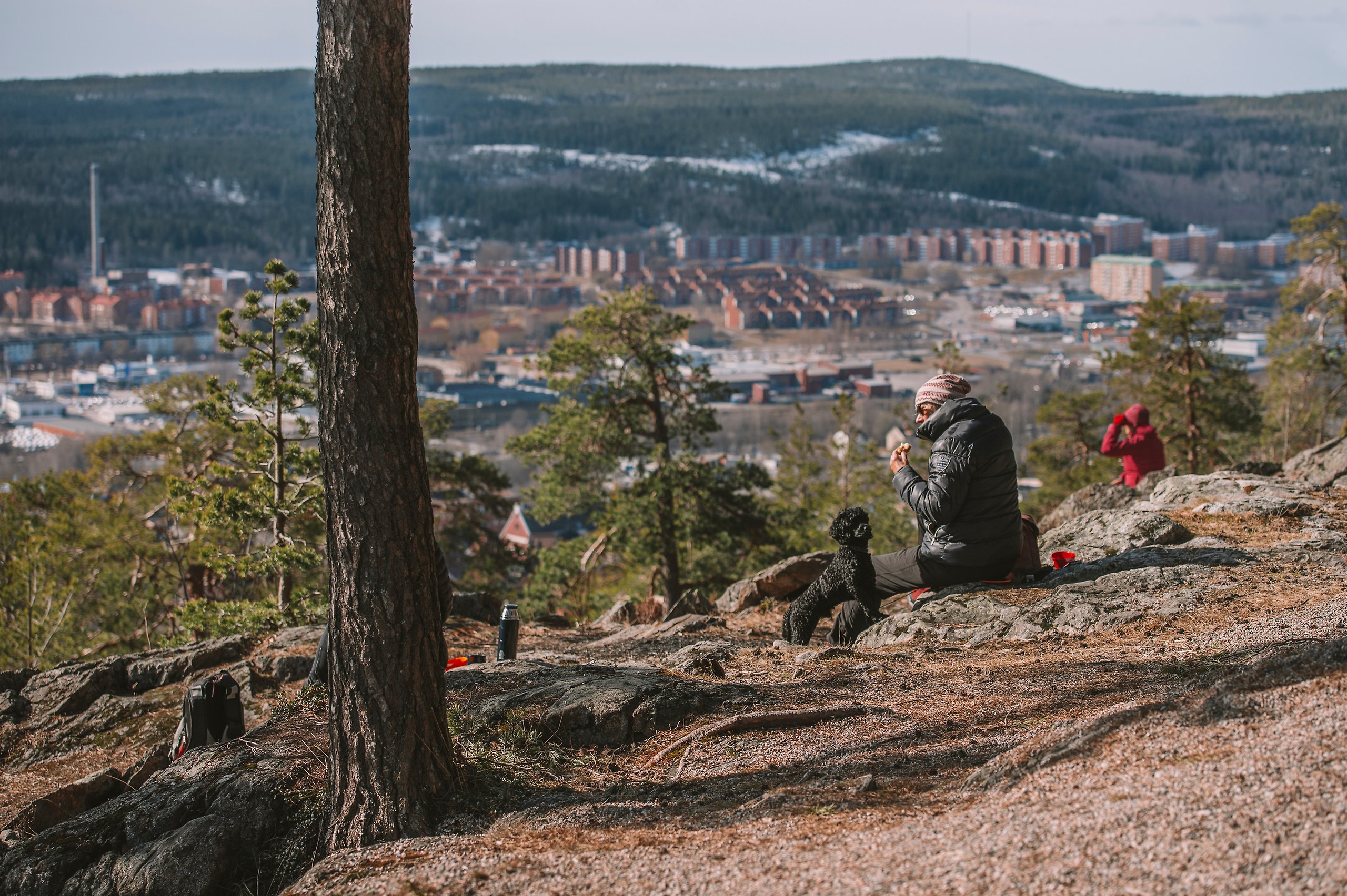 Två personer, en med svart jacka och en med röd jacka sitter på stenarna vid Norra Berget i Sundsvall. De har en svart hund med sig också. Framför dem breder Stenstan ut sig i sin fulla prakt. En fantastisk utsikt över stad, hav, berg och land.